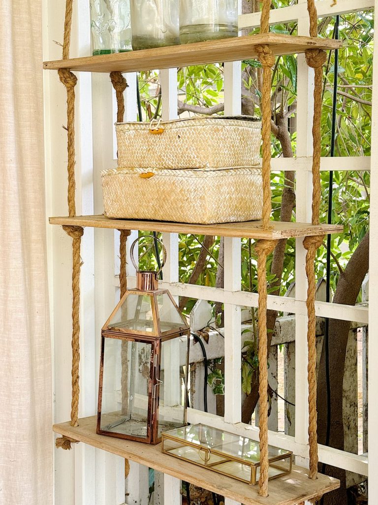 A rustic wooden shelf with wicker baskets, glass jars, and a lantern, located by a window with sheer curtains.