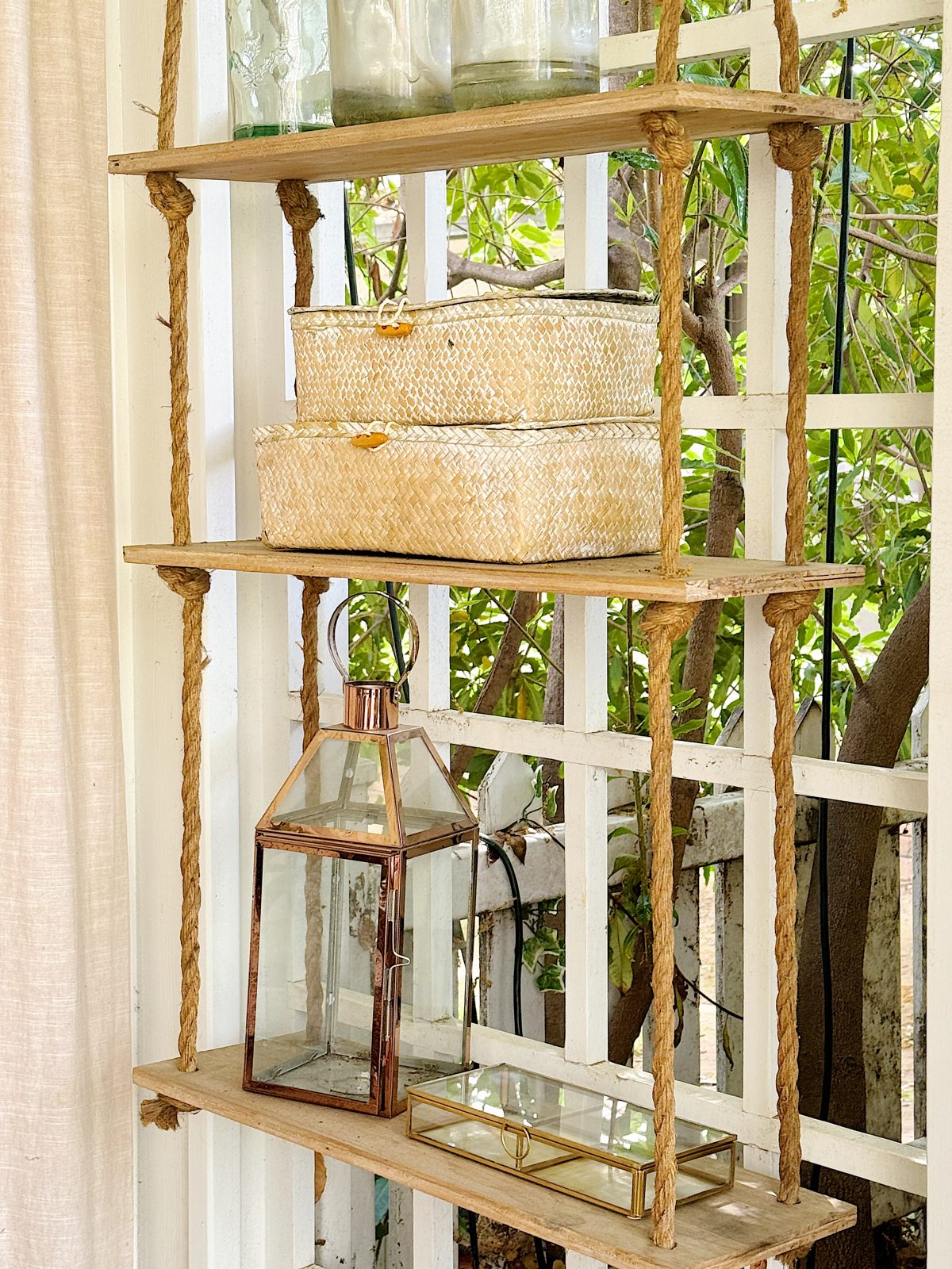 A rustic wooden shelf with wicker baskets, glass jars, and a lantern, located by a window with sheer curtains.
