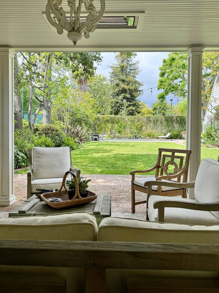 View from an enclosed patio with wicker furniture overlooking a lush garden with green lawn and trees under a cloudy sky.