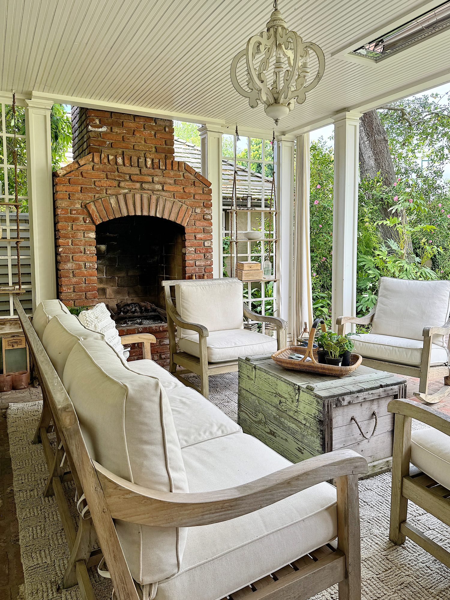 Cozy enclosed porch with brick fireplace, wooden furniture with white cushions, and a rustic storage chest, surrounded by lush greenery visible through large windows.