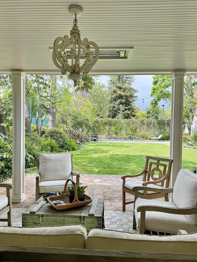 A covered patio furnished with a white sofa, two chairs, and a coffee table, overlooking a lush garden through large open archways.