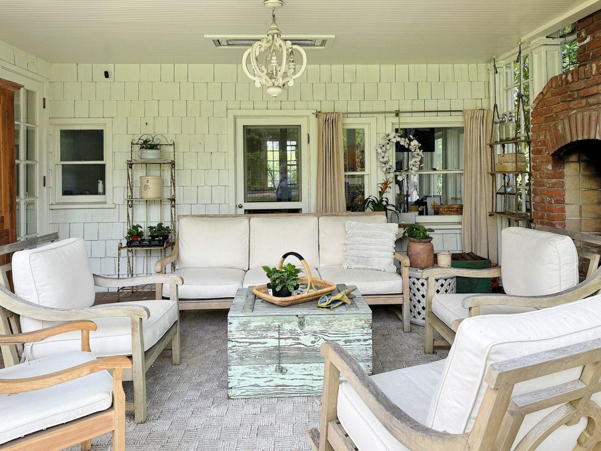 A cozy sunroom with white furniture, a brick fireplace, wooden accents, and decorative plants, illuminated by natural light from surrounding windows.