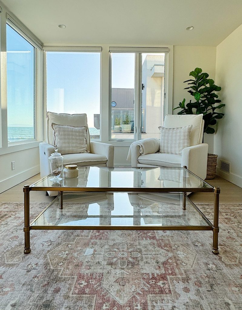 Bright living room with two white armchairs, a glass coffee table, and a large window overlooking the ocean.