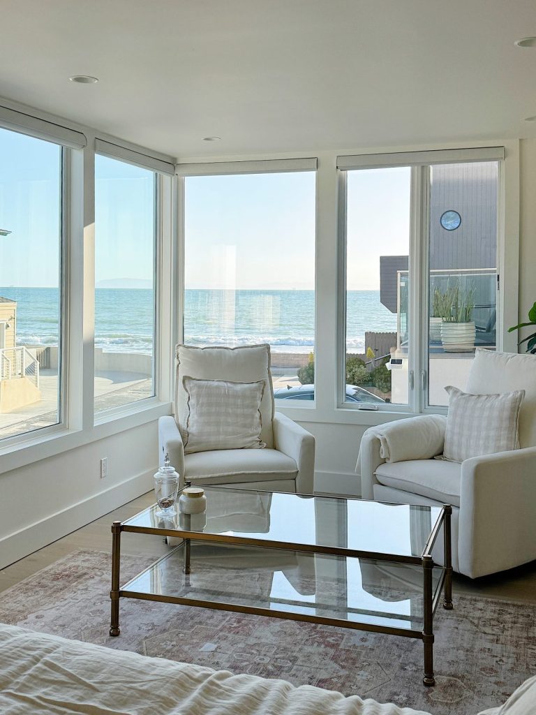 Bright, modern living room with two white armchairs, a glass coffee table, and large windows overlooking a beach and ocean.