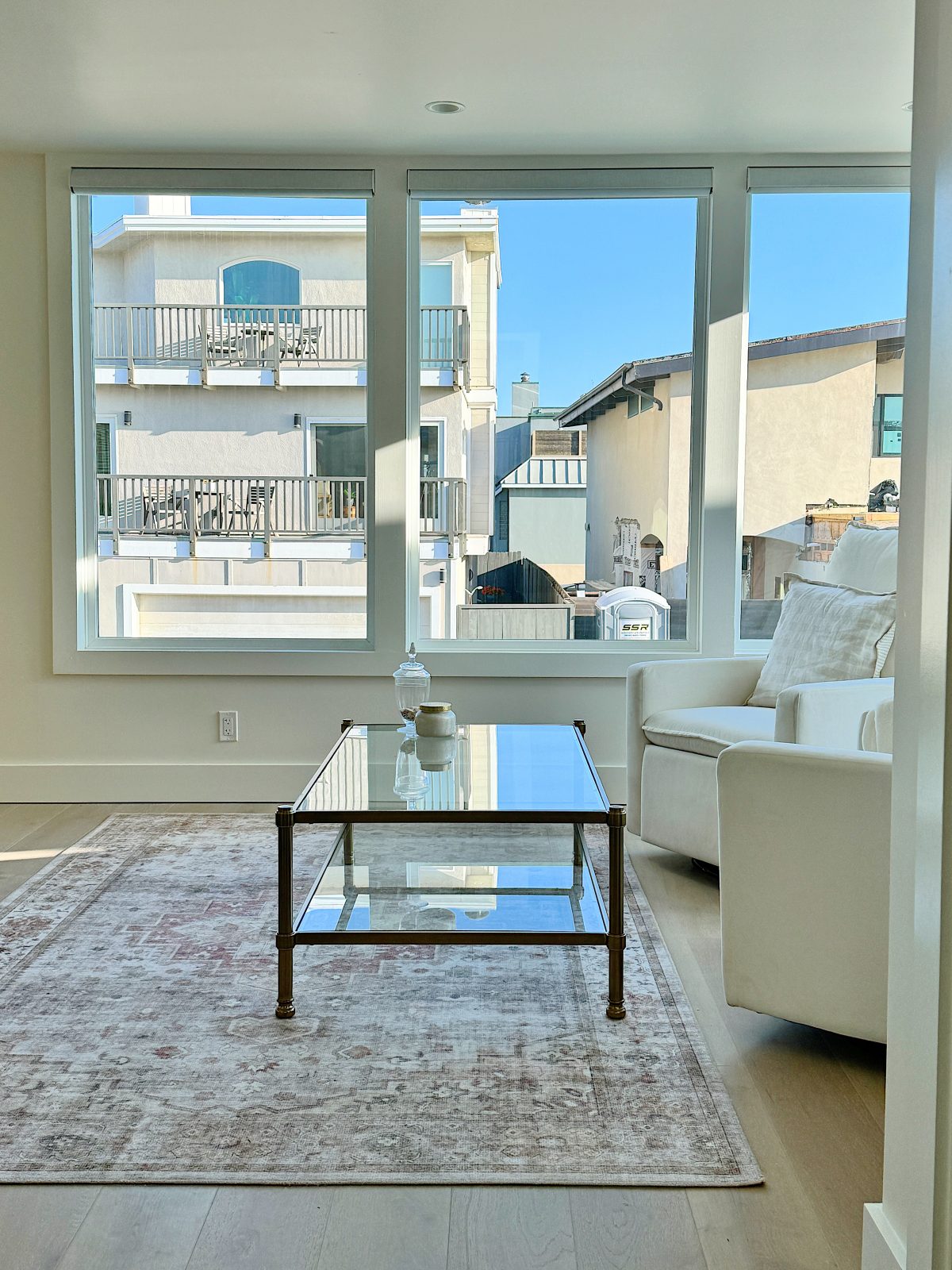 Modern living room with a glass coffee table, white sofa, and large windows showing neighboring houses.
