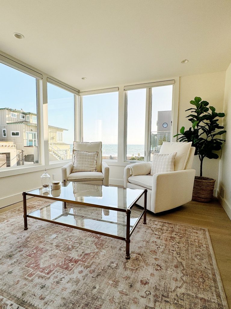 Bright living room with large windows overlooking the beach, featuring two white chairs, a glass coffee table, and a potted plant.