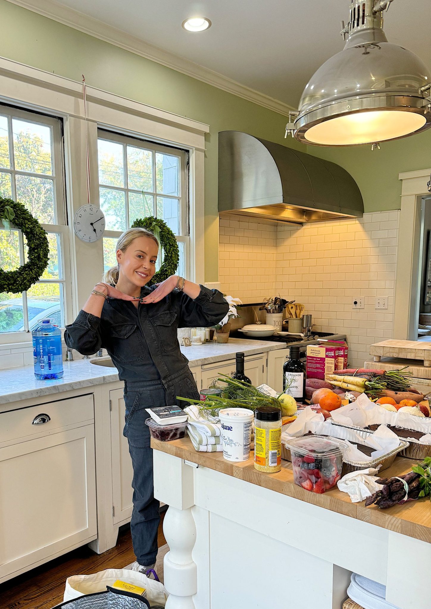 A woman smiles while standing in a kitchen with fruits and vegetables spread out on the counter.