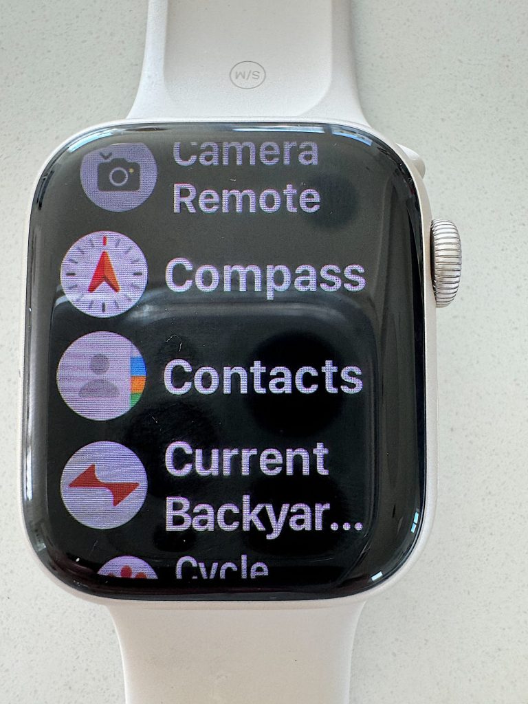 Close-up of a smartwatch displaying various apps including camera remote, compass, contacts, and current backyard.