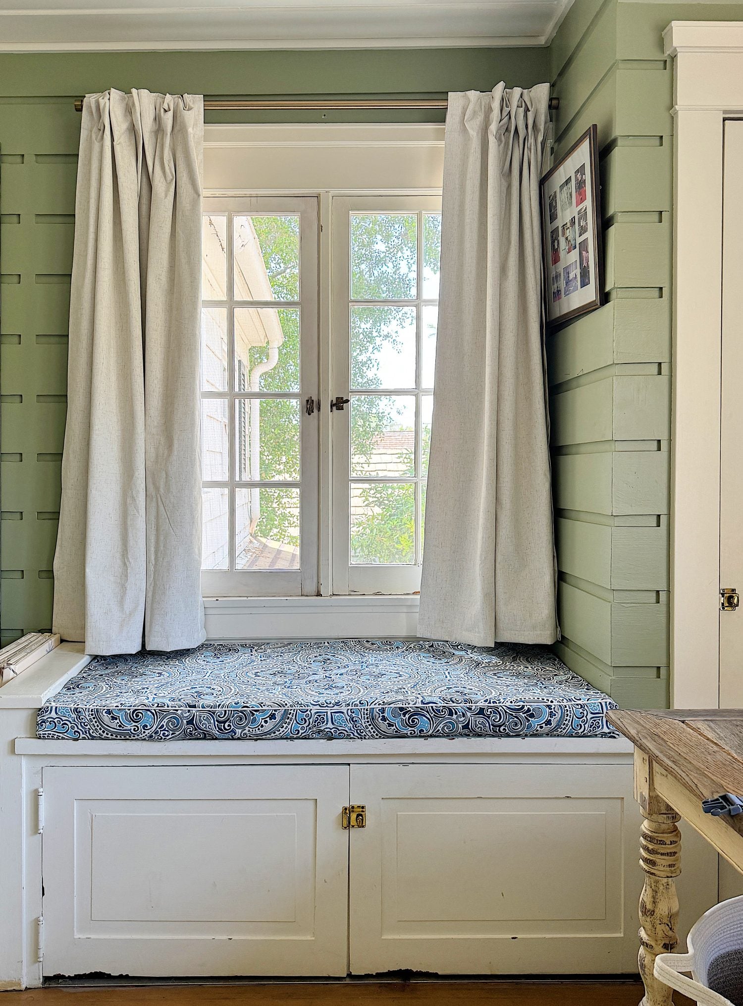 A cozy nursery with a white crib on the right, a rustic wooden table with baskets underneath by a window, and soft green walls.