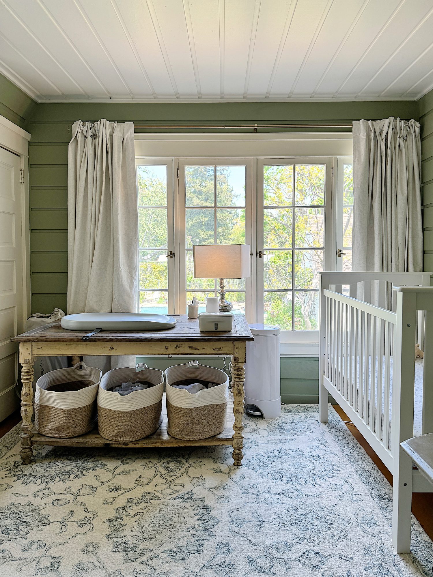 A cozy nursery with a white crib on the right, a rustic wooden table with baskets underneath by a window, and soft green walls.