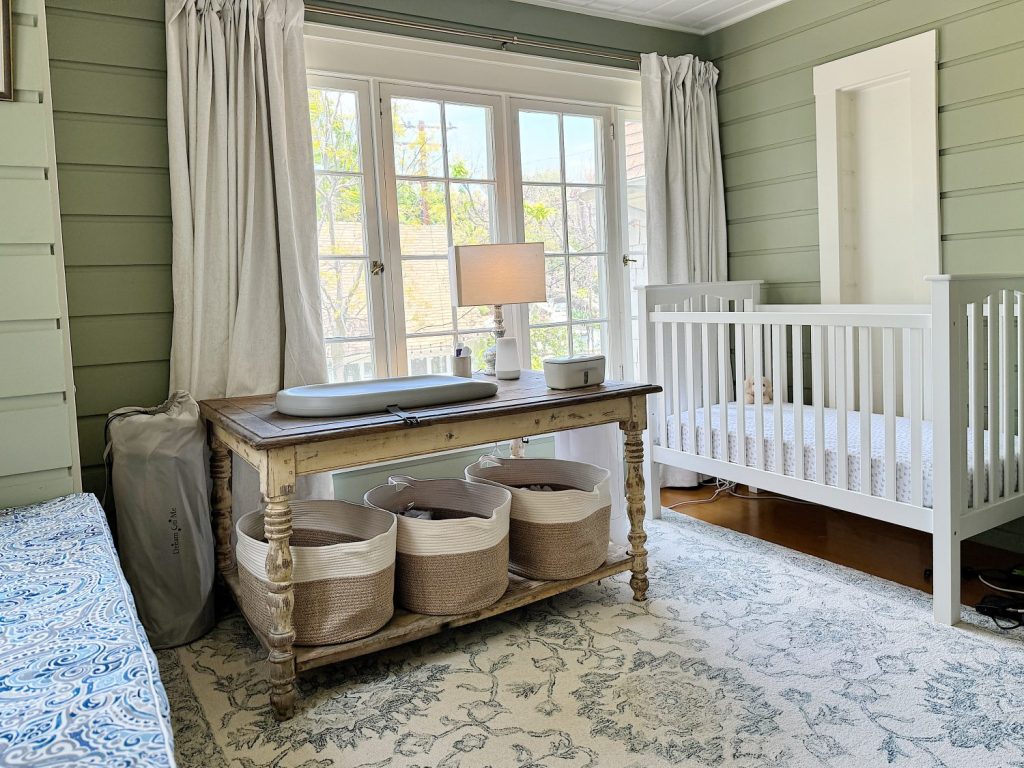 A cozy nursery with a white crib on the right, a rustic wooden table with baskets underneath by a window, and soft green walls.