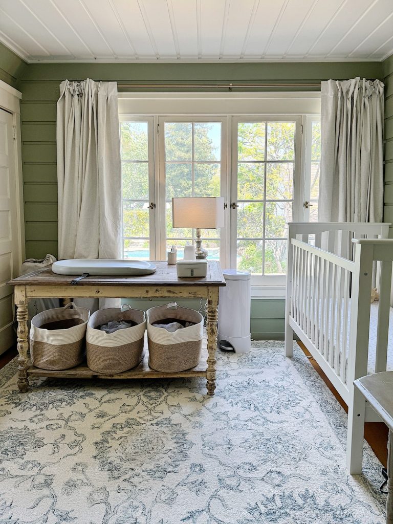 A cozy nursery with a white crib on the right, a rustic wooden table with baskets underneath by a window, and soft green walls.