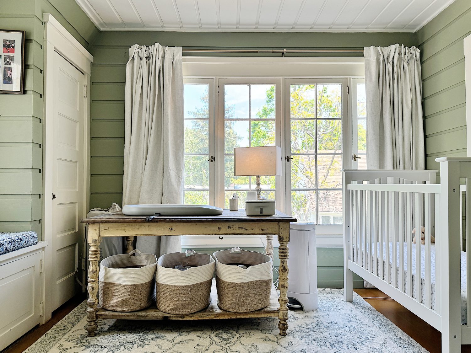 A cozy nursery with a white crib on the right, a rustic wooden table with baskets underneath by a window, and soft green walls.