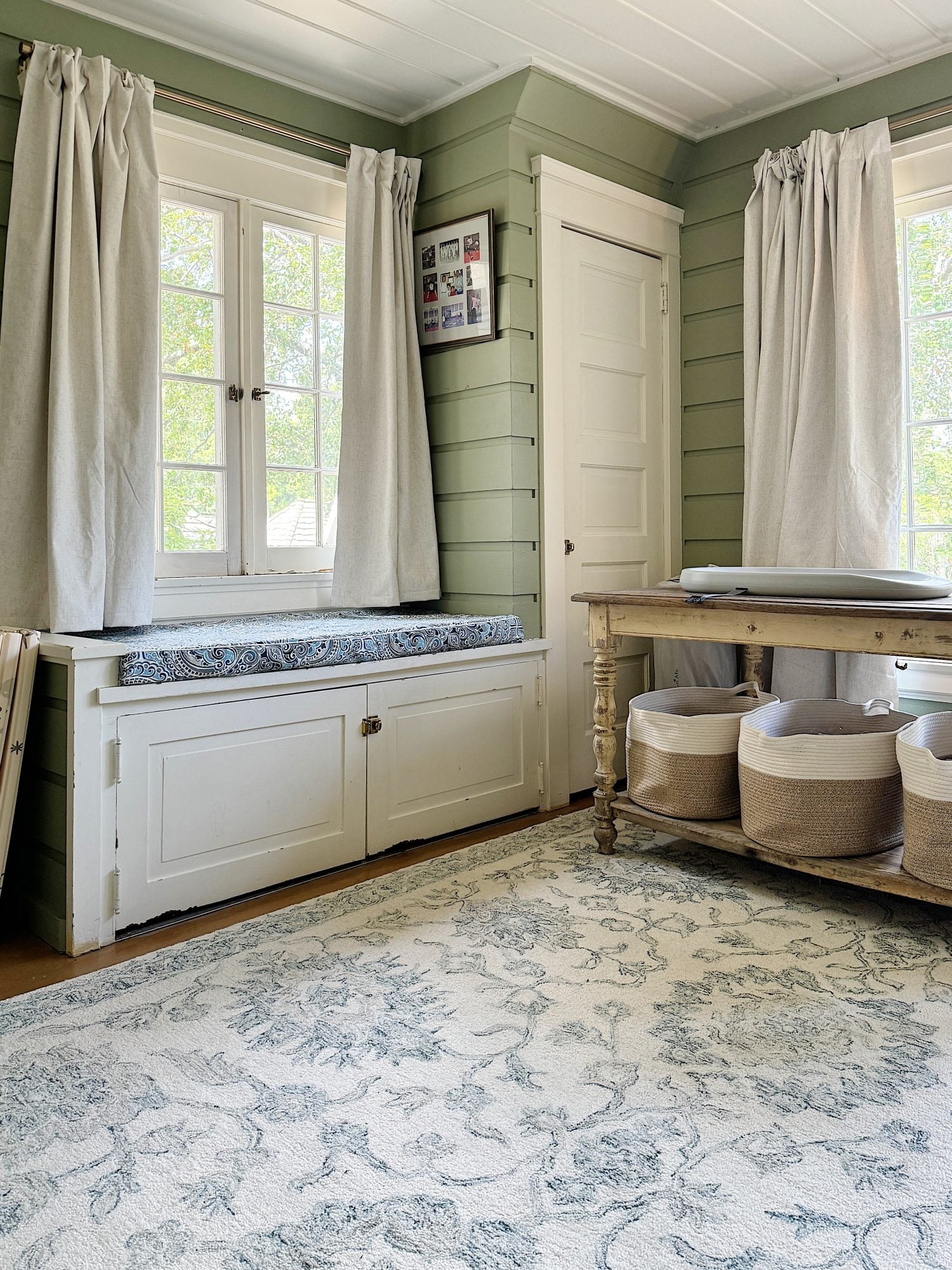 A cozy nursery with a white crib on the right, a rustic wooden table with baskets underneath by a window, and soft green walls.