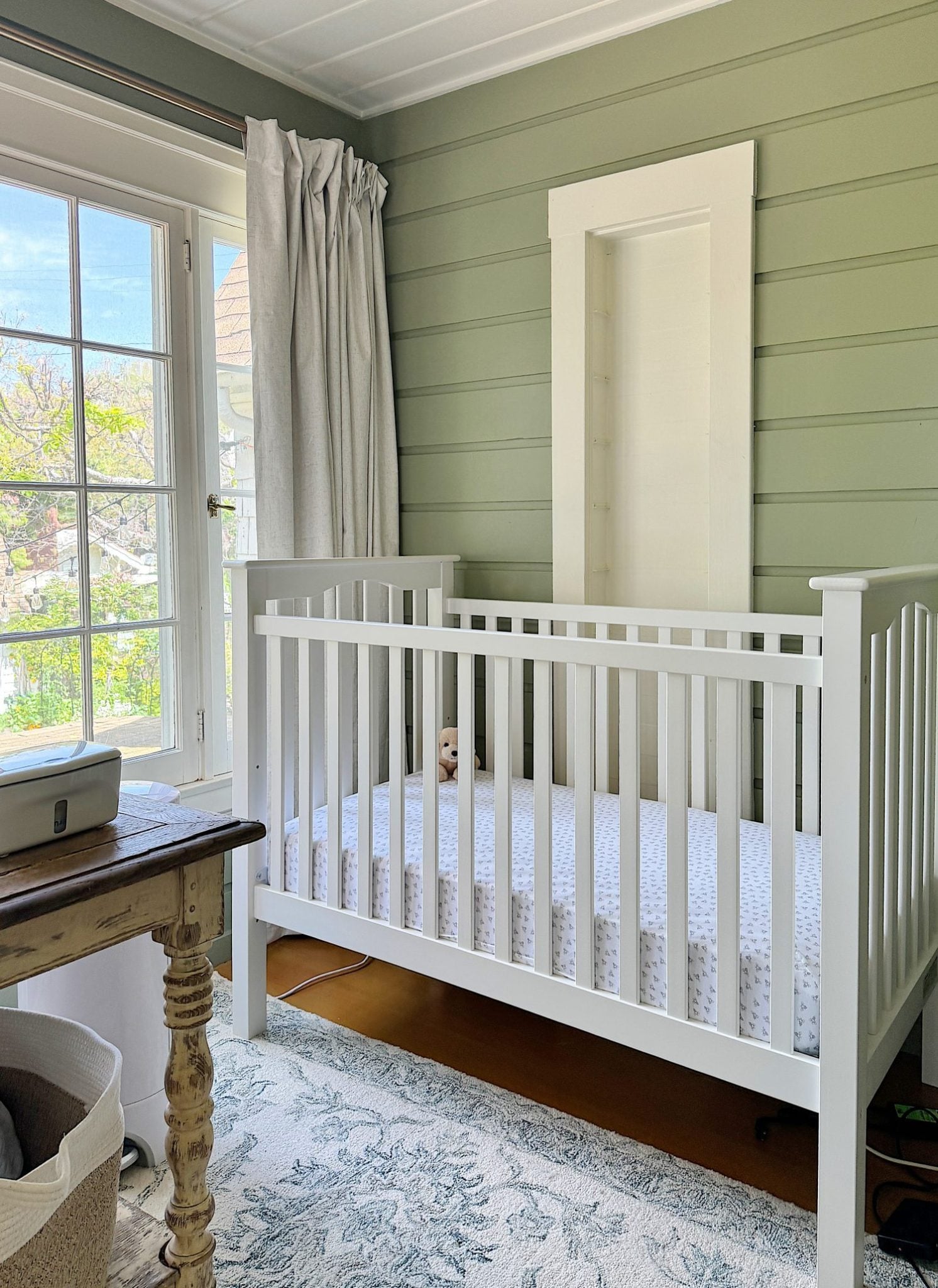 A cozy nursery with a white crib on the right, a rustic wooden table with baskets underneath by a window, and soft green walls.