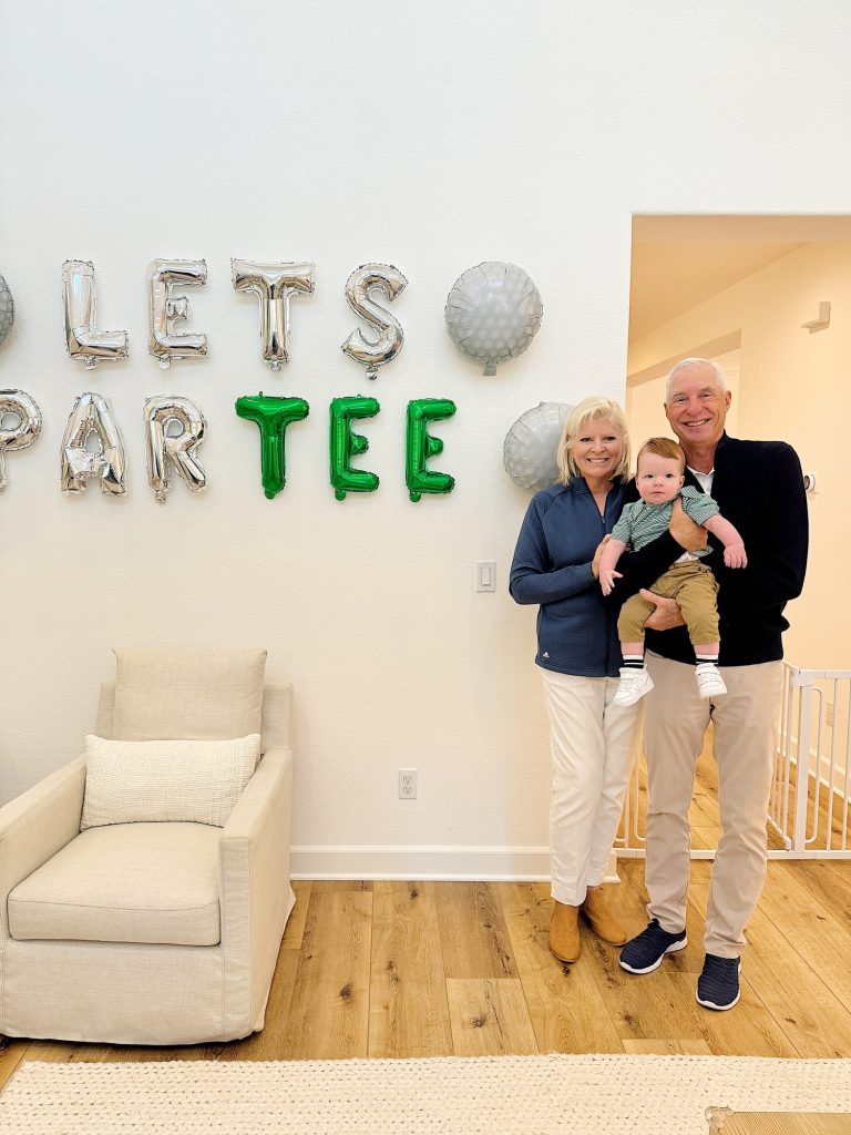 A family moment with grandparents holding a toddler, standing in a room decorated with a playful "let's par tee" golf-themed wall decor.