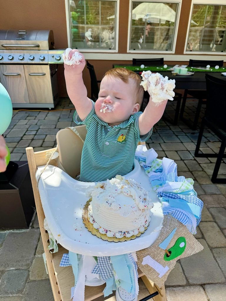 A baby sitting in a high chair celebrating with a cake, with frosting covering their hands.
