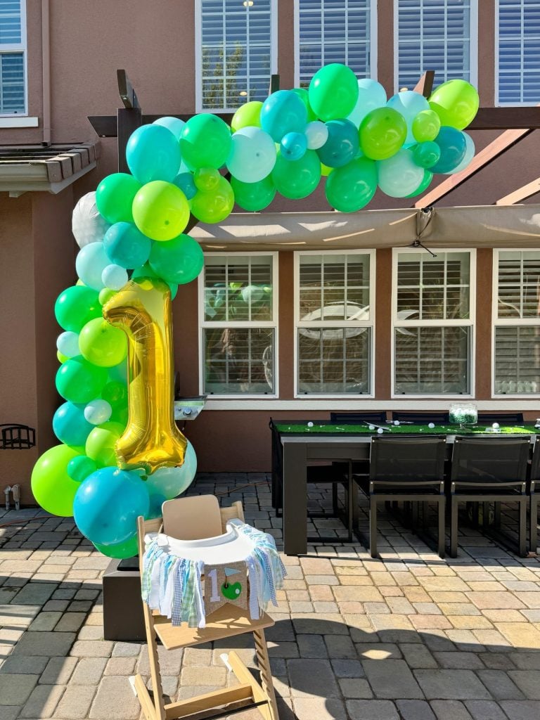 Outdoor first birthday celebration setup with a high chair and a balloon arch in shades of green and blue.