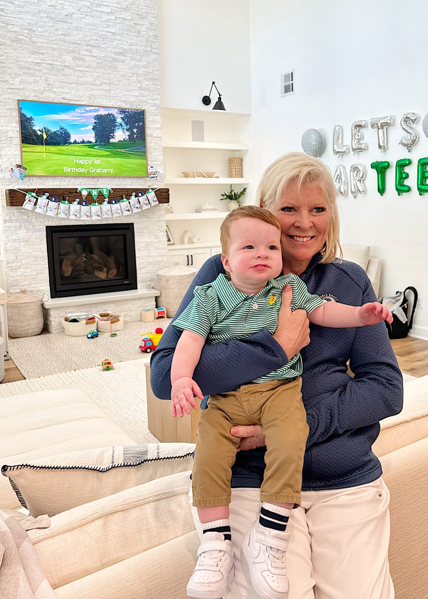 A smiling woman holding a baby in a living room decorated for a birthday party.