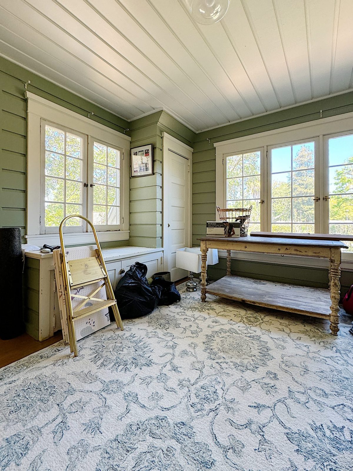 A bright, spacious room with large windows, furnished with a wooden table, built-in bench bench, and a patterned rug.