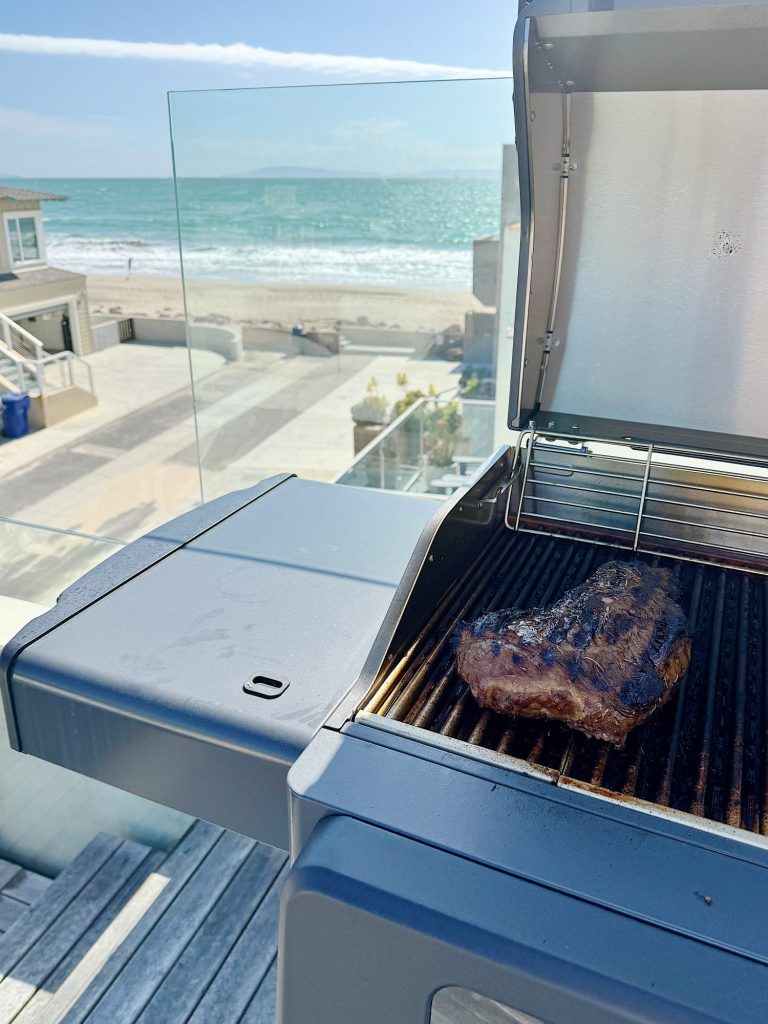 A steak grilling on a barbecue overlooking a beachfront view with a clear sky and calm sea.
