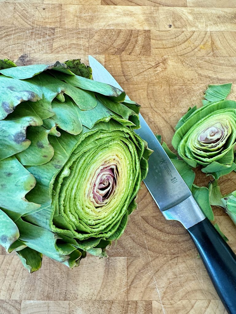 A halved artichoke next to a whole artichoke and a chef's knife on a wooden cutting board.