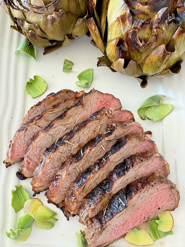 Grilled steak sliced and served with roasted artichokes on a white plate, garnished with herbs.
