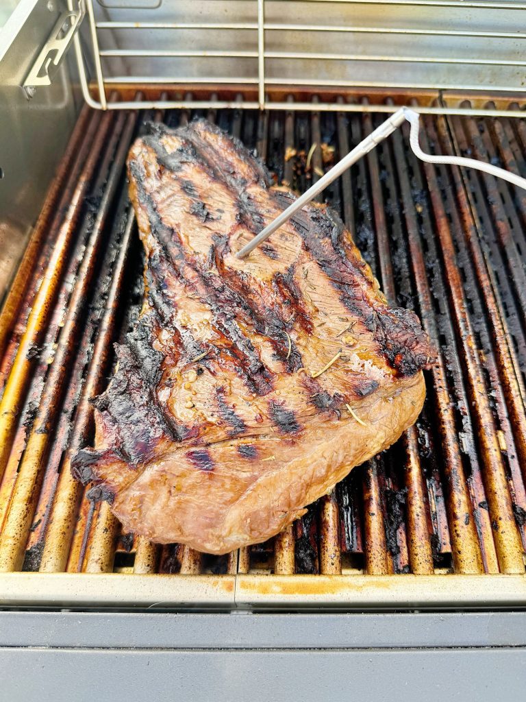 A large steak with grill marks and a meat thermometer inserted, cooking on a gas grill.
