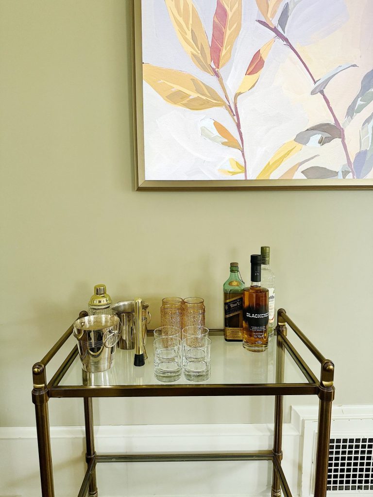 A small bar cart with bottles of liquor, glasses, and an ice bucket, positioned under a large botanical painting in a room with cream walls.