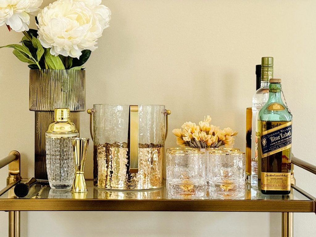 A home bar setup on a table displaying a vase of white flowers, cocktail-making equipment, and bottles of liquor including johnnie walker blue label.