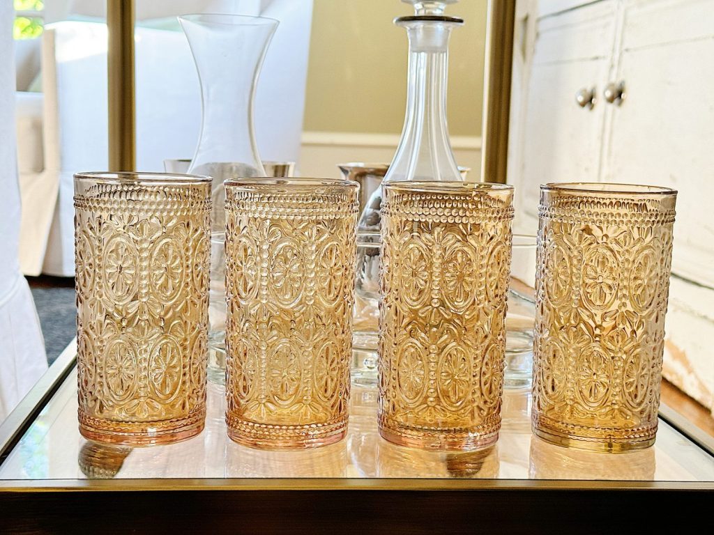 Four ornate golden glass tumblers on a metal tray, with a decanter in the background on a wooden table.