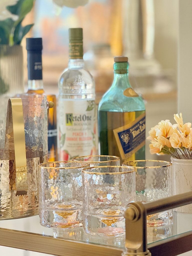 Assortment of liquor bottles and elegant, textured glass tumblers on a gold bar cart, with soft-focus background.