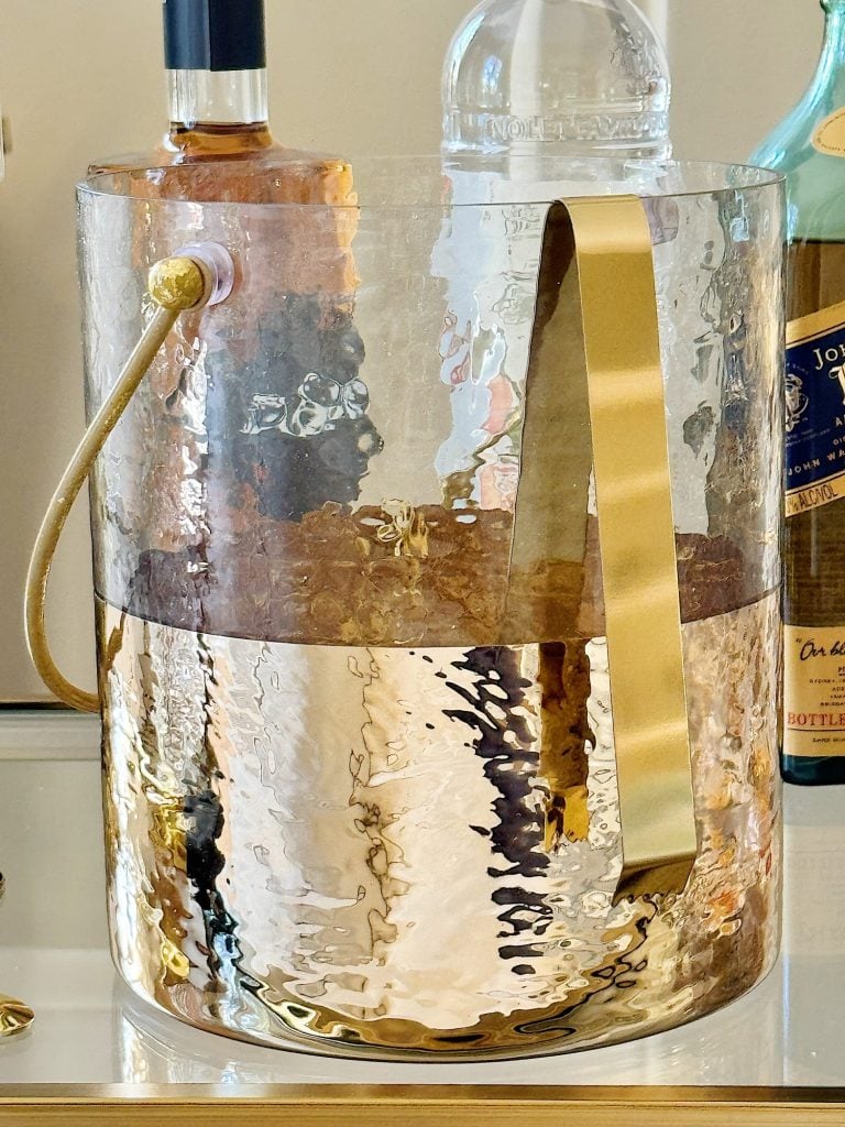 A clear glass pitcher filled with a golden liquid, featuring a brass handle and a blurred background with bottles.