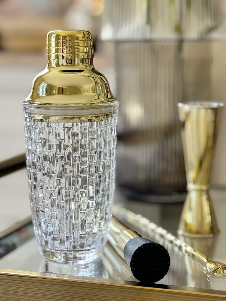 A golden cocktail shaker set on a glass tray, featuring a textured glass body and metallic top, with a jigger and a bar spoon beside it.