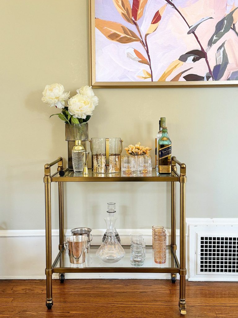 A stylish bar cart with two shelves, decorated with flowers, glassware, and liquor bottles, in a room with beige walls.