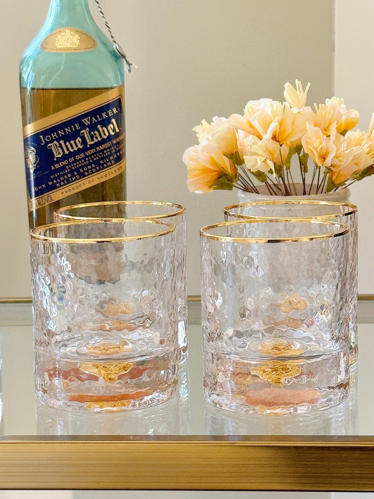 A stylish bar cart with two shelves, decorated with flowers, glassware, and liquor bottles, in a room with beige walls.