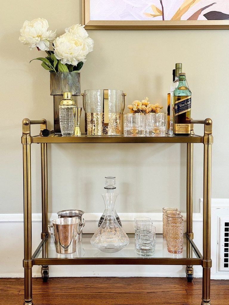 A stylish bar cart with two shelves, decorated with flowers, glassware, and liquor bottles, in a room with beige walls.