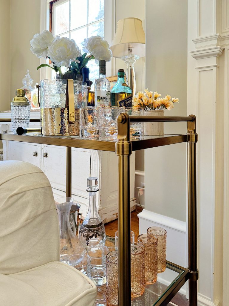 Elegant bar cart with alcoholic beverages, decorative glasses, and floral arrangements in a cozy, well-lit room.