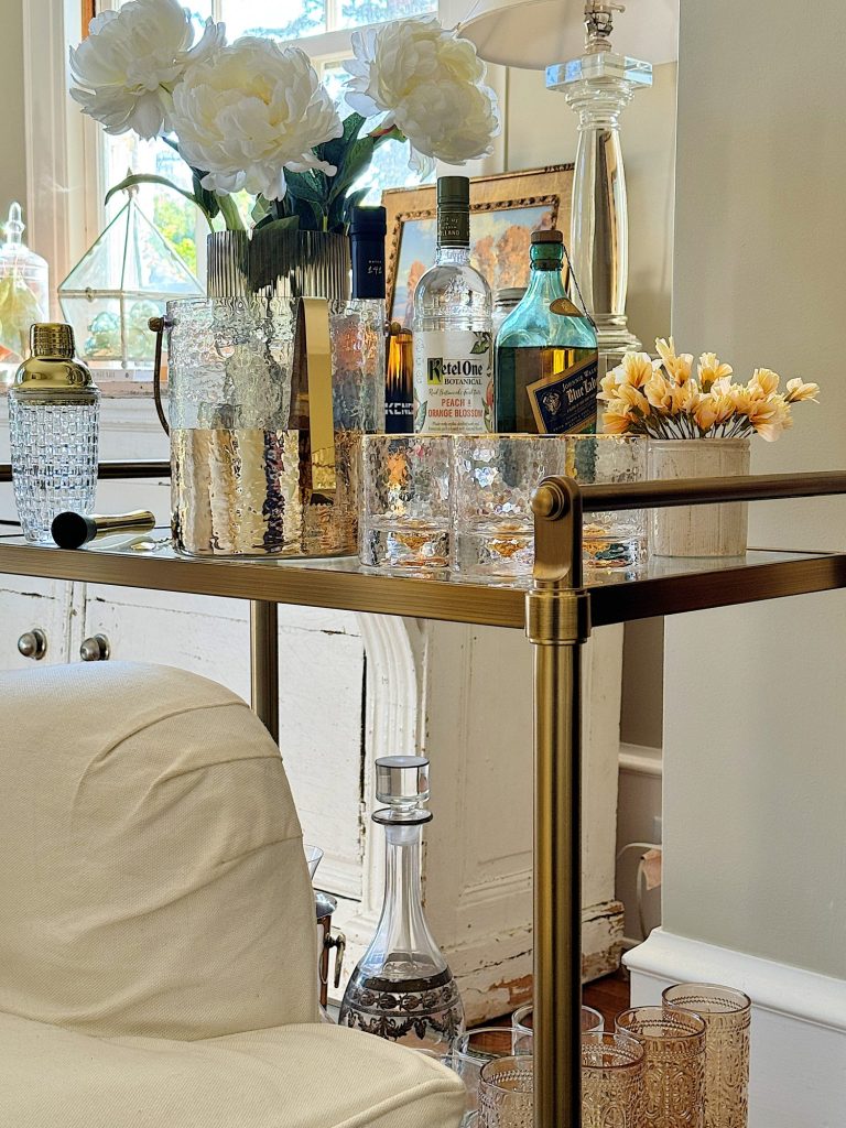 A well-appointed bar cart with various liquor bottles, glassware, and floral decorations in a cozy room setting.