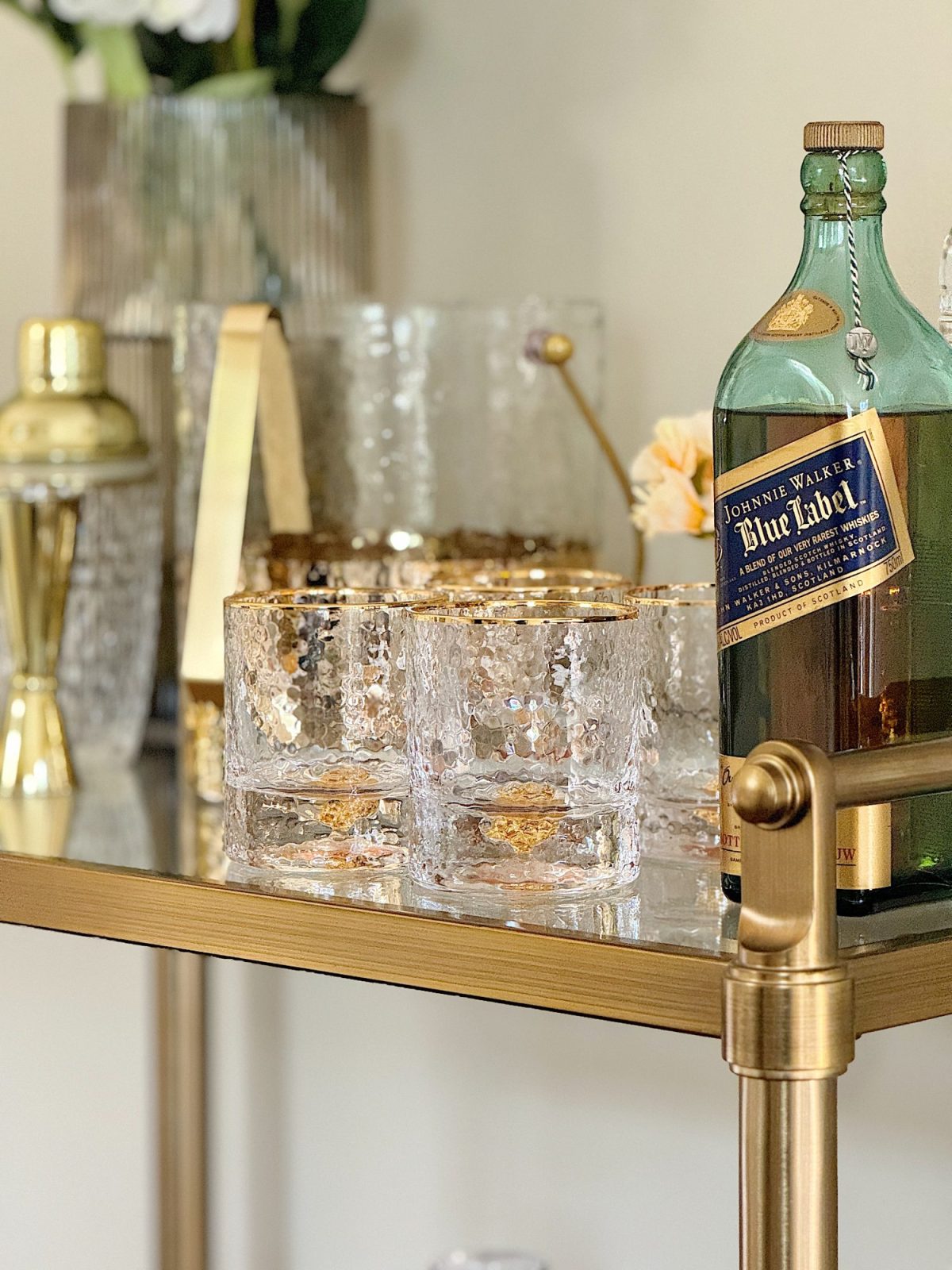 A gold bar cart displaying a bottle of johnnie walker blue label, glass tumblers with a textured design, and decorative items in a home setting.