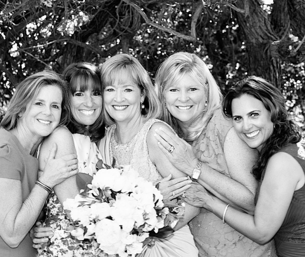 Five smiling women in elegant dresses pose together holding a bouquet, standing under a tree with dappled sunlight.