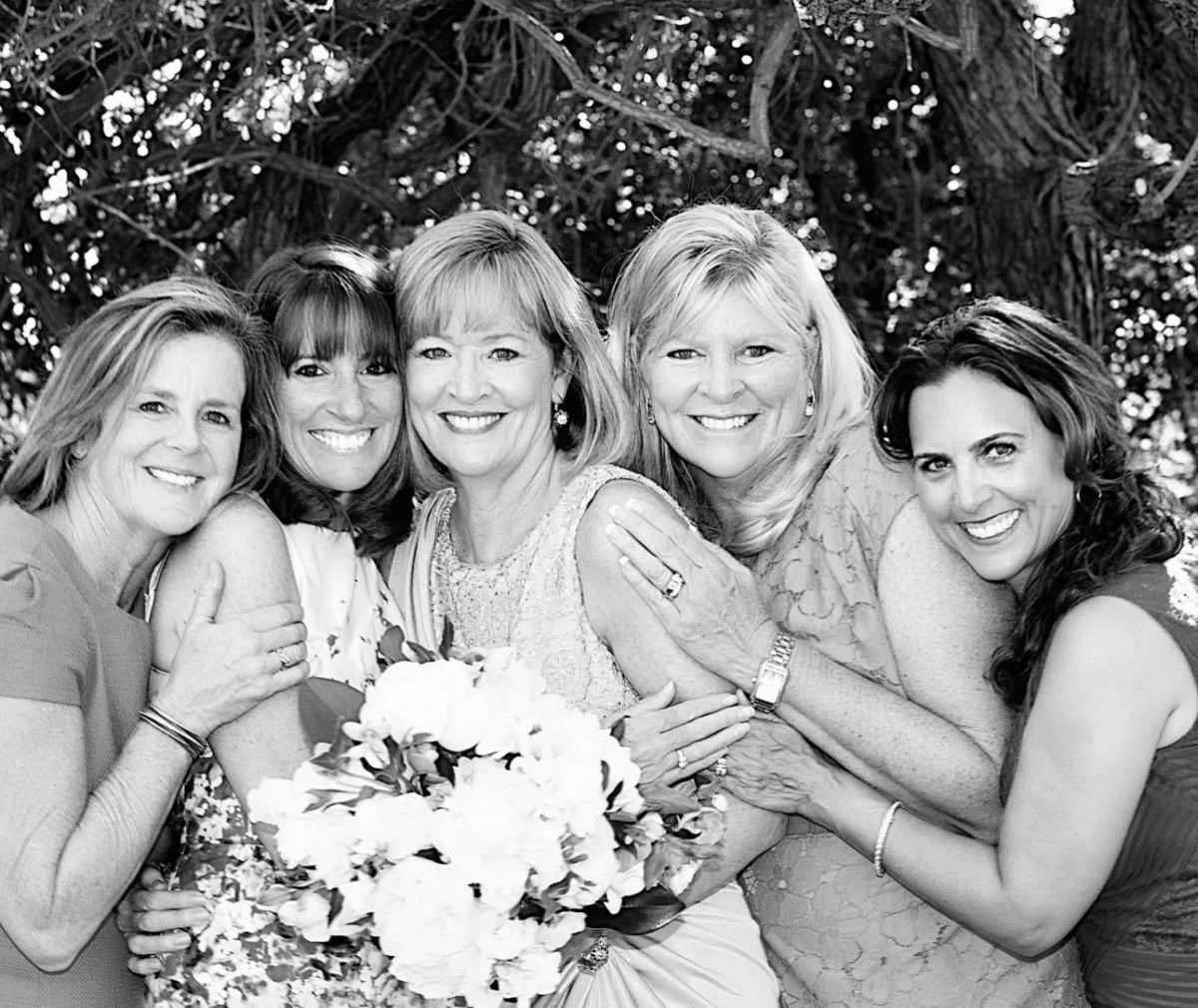 Five smiling women in elegant dresses pose together holding a bouquet, standing under a tree with dappled sunlight.