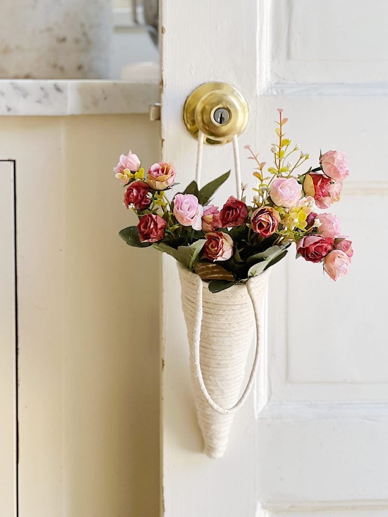 Door with a decorative knitted hanging vase filled with colorful roses, mounted on a white wooden surface with a gold handle.
