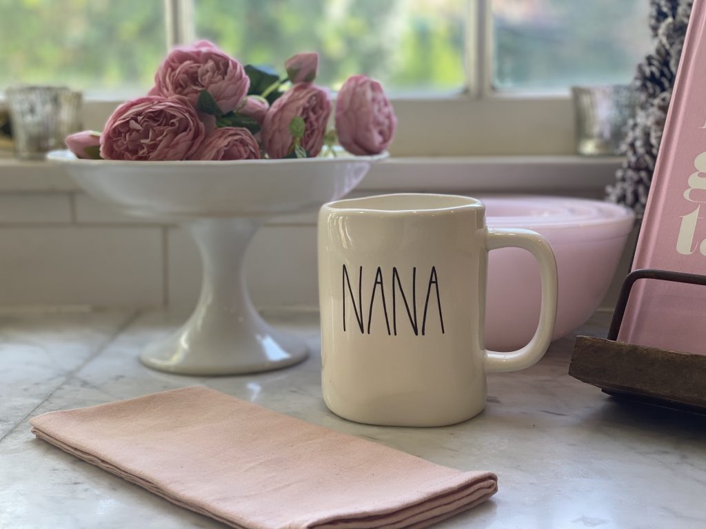 A mug with the word "nana" printed on it, placed on a kitchen counter beside a bouquet of pink roses, often seen during family gatherings.