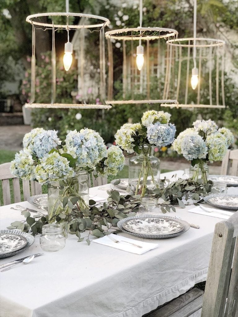 An outdoor dining setup with rustic wooden table, floral arrangements, and hanging light fixtures.