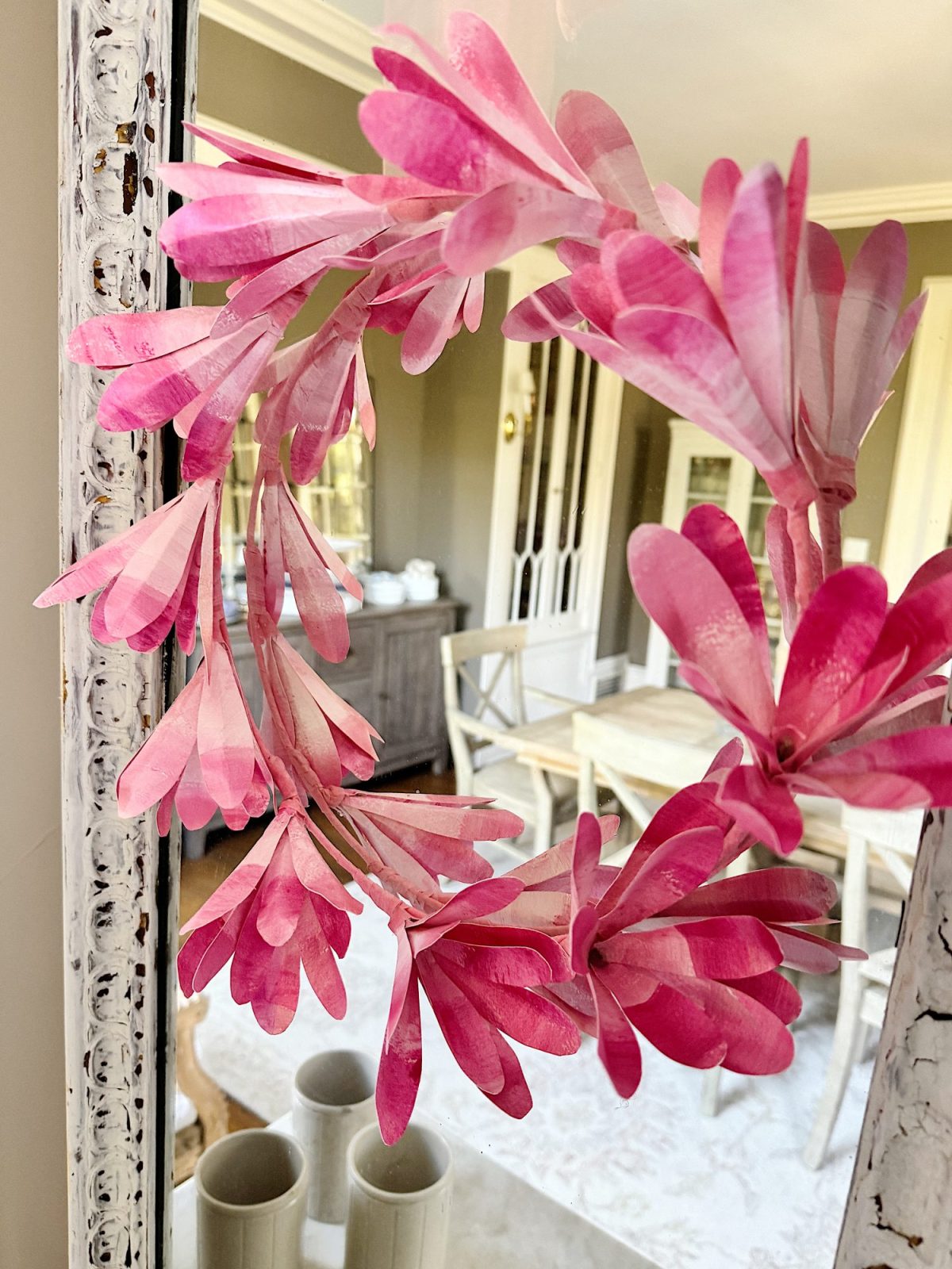 Pink paper DIY wreath hanging on a distressed white mirror frame with a blurred interior background.