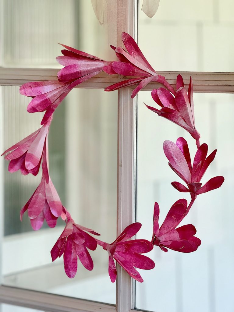 A pink decorative wreath made of paper hanging in front of a window.