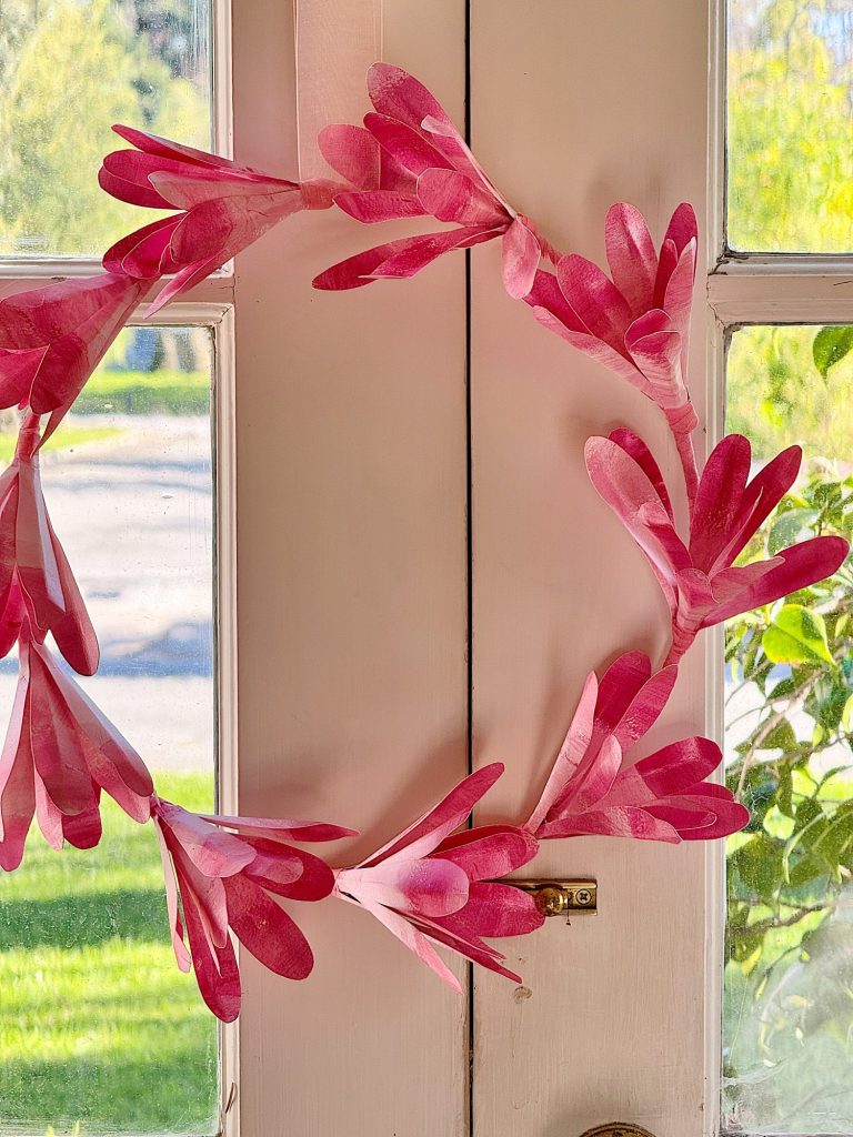 A pink paper DIY wreath hanging on a white door with a window, allowing sunlight to filter through.