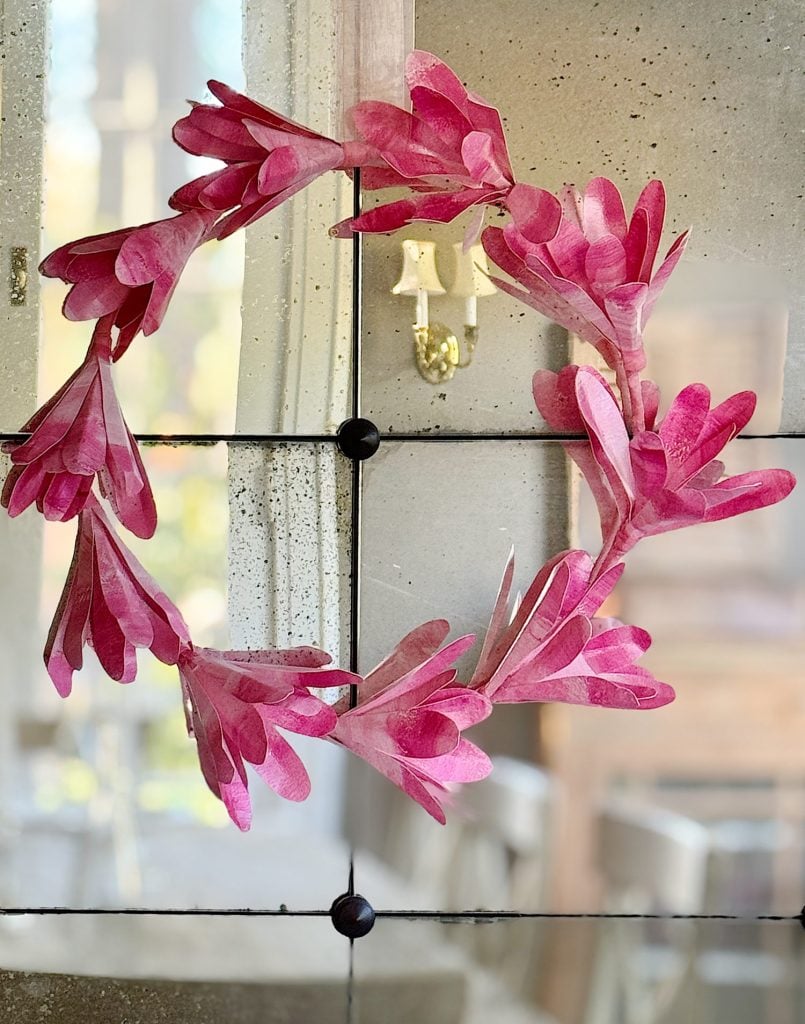 A pink floral paper wreath hanging on a glass door with a blurred chandelier in the background.
