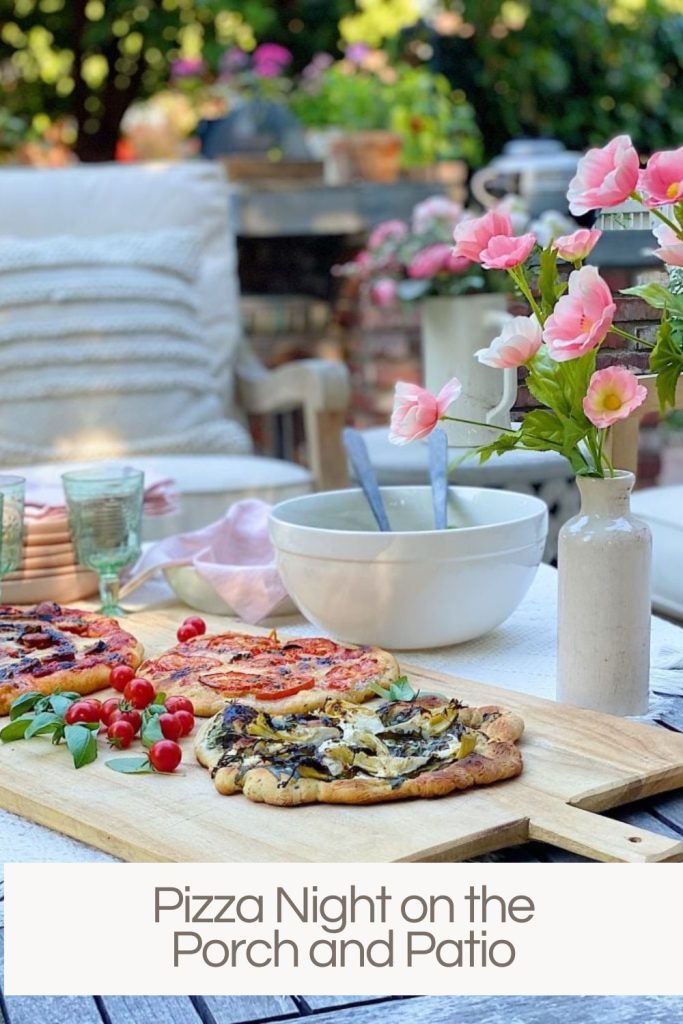 Table with homemade pizzas, a bowl of salad, and a vase of pink flowers on an outdoor porch with patio furniture in the background.
